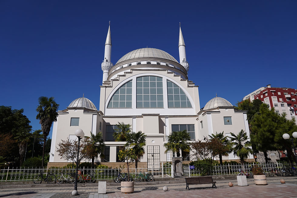 White mosque with two minarets under a clear blue sky, surrounded by trees and benches. Bicycles parked by white fence in foreground.