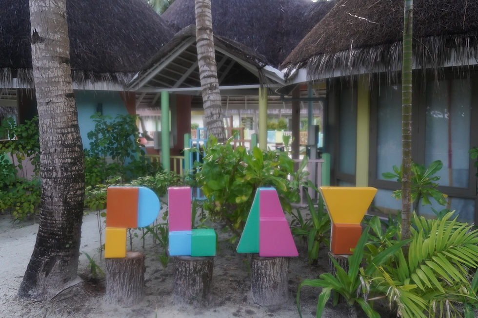 Colorful letters spelling "PLAY" on tree stumps, surrounded by plants and palm trees. Background has thatched-roof hut and pastel shades. Tropical vibe.