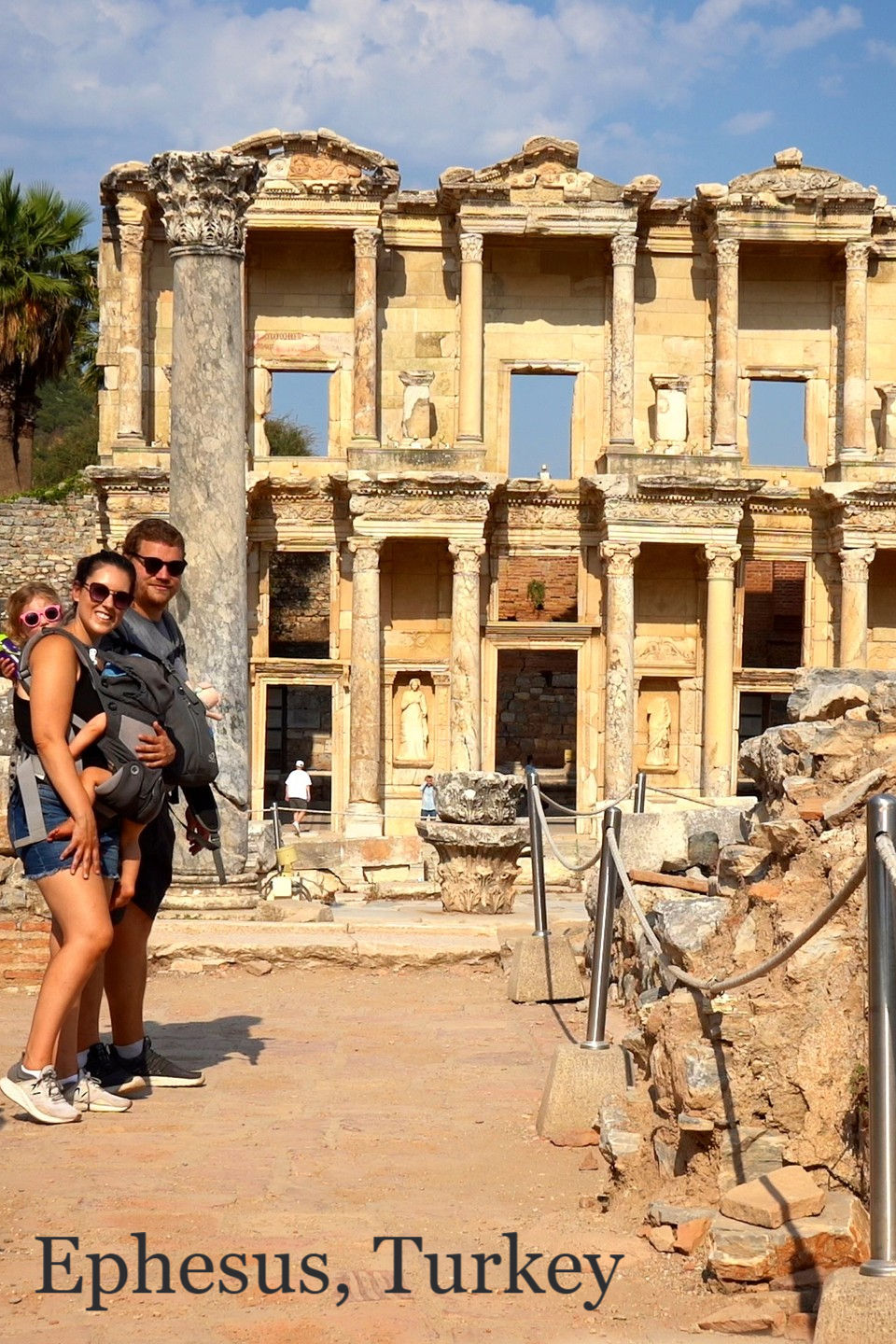 Family with toddler in carrier smiles in front of ancient Celsus Library ruins, Ephesus, Turkey. Clear blue sky and historic stonework.