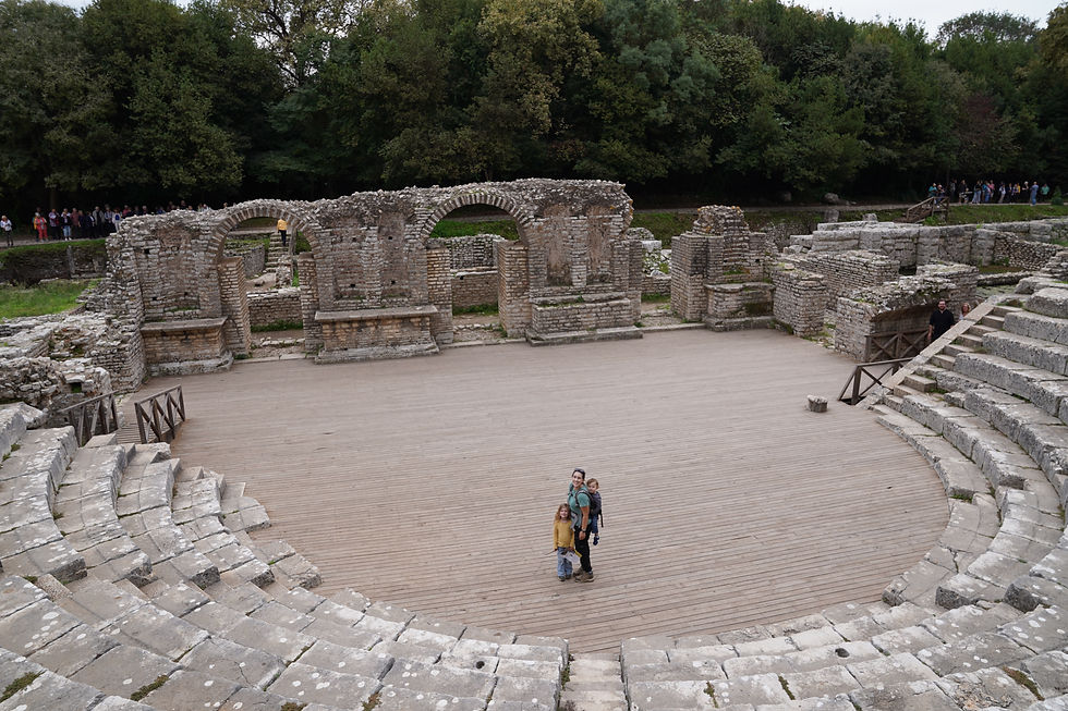 A family stands on an ancient amphitheater stage, surrounded by stone ruins and green trees, with a small crowd in the background.