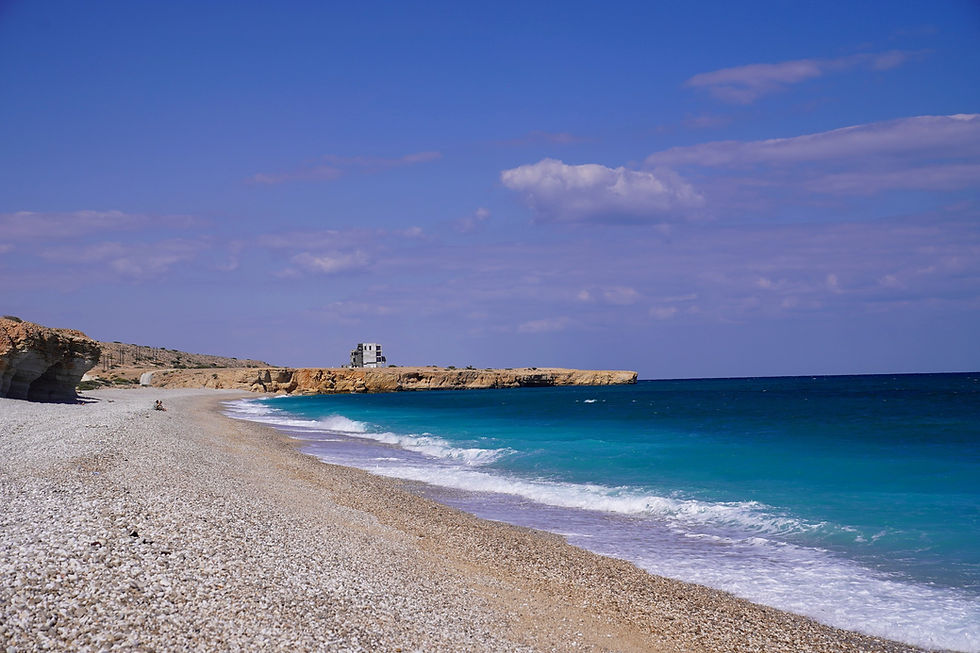 Remote beach with turquoise waves, pebbled shore, and rocky cliffs under a bright blue sky. Lonely structure on the horizon.