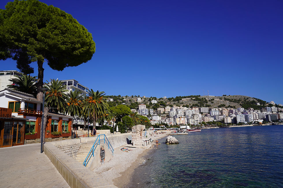 Coastal cityscape with a clear blue sky, palm-lined promenade, and calm sea. People relax on the pebble beach. Hillside buildings in view.