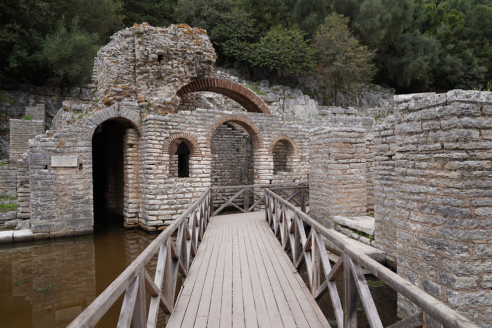 Stone ruins with arches stand by a wooden walkway over water. Lush green trees surround the scene, creating a serene atmosphere.