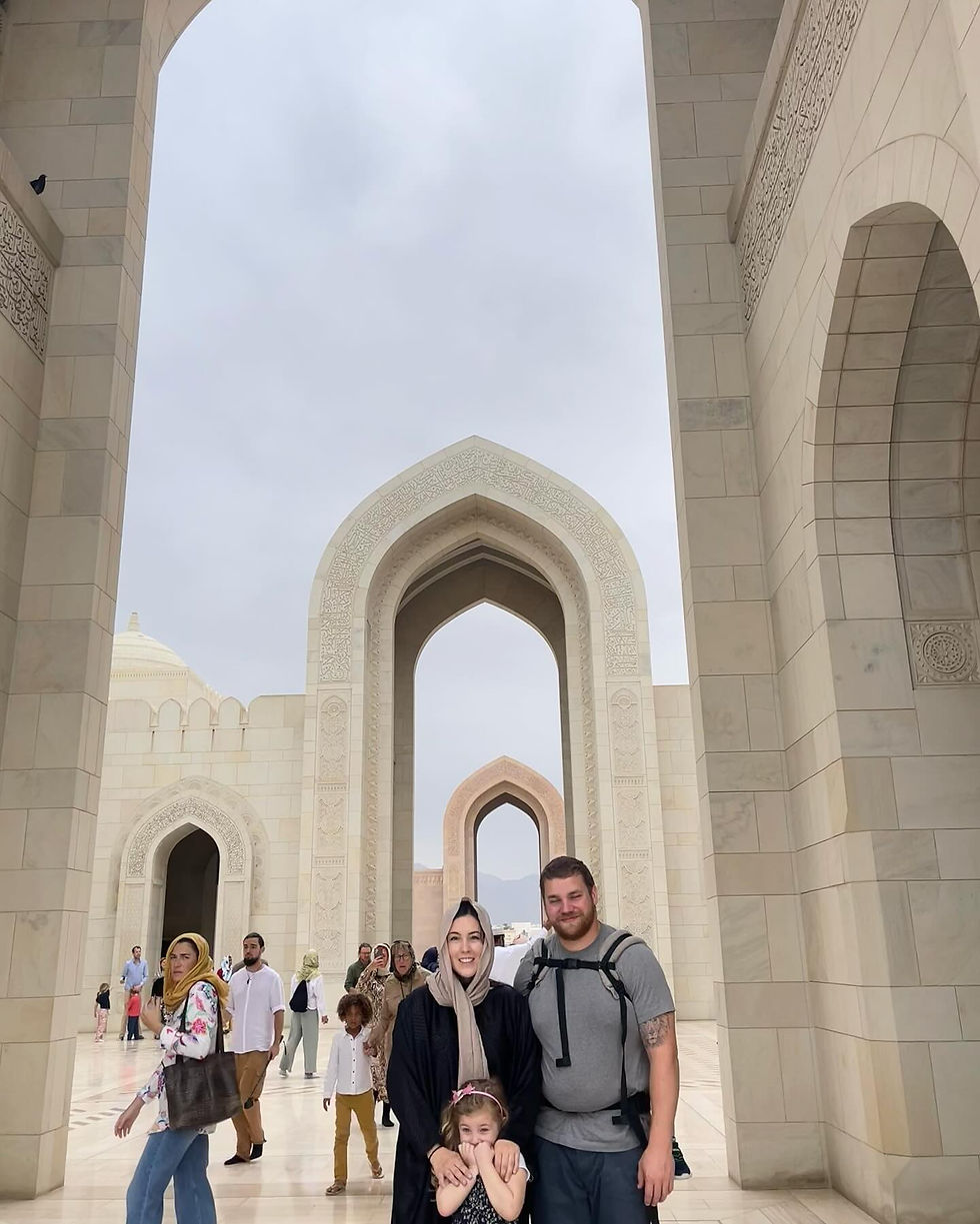 A smiling family poses in front of ornate archways at a mosque. Other visitors walk around. The sky is overcast, creating a serene mood. The location is Sultan Grand Qaboos Mosque in Muscat, Oman.