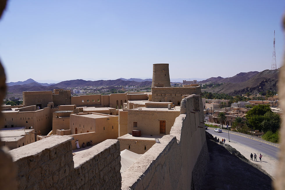 View of a historic desert town with tan buildings and a fortress tower. Mountains and clear blue sky in the background; people walking below.