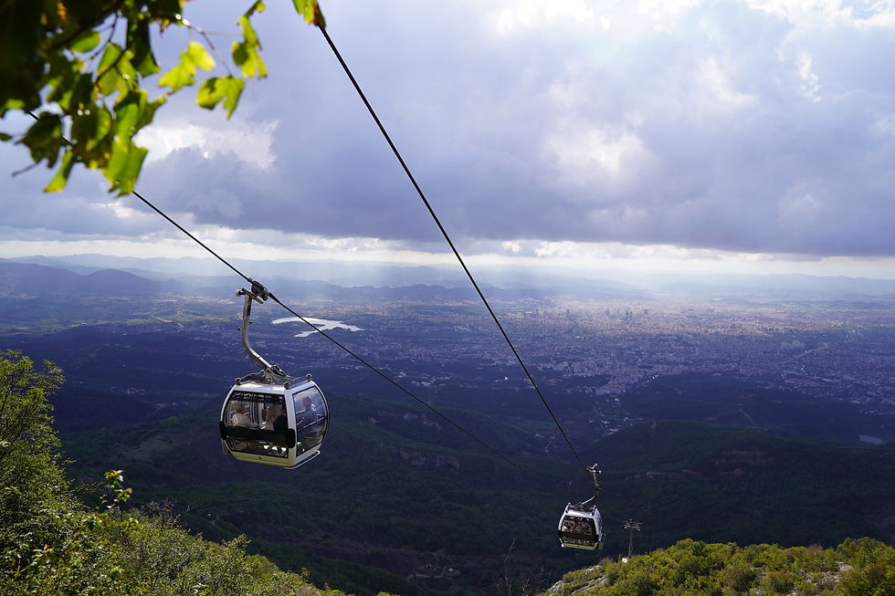 Cable cars glide over a lush green landscape with a city below. The sky is cloudy, with rays of sunlight breaking through.