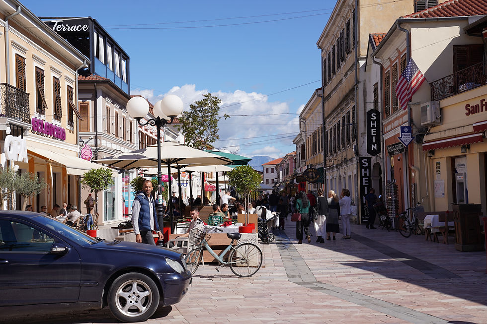 Bustling street with people walking, sitting at cafes. Blue car, bicycle, and umbrellas in foreground. Bright day, shops with signs like "Shein".