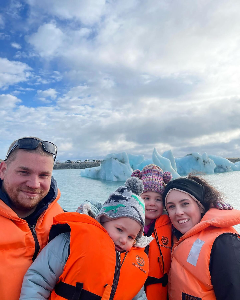 Family in orange life jackets smiles in front of icy blue glaciers under a cloudy sky. Two kids wear colorful pom-pom hats, creating a joyful mood.