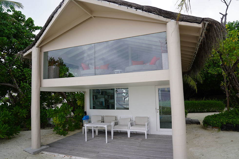 Two-story beach hut with glass upper level. White chairs, table on wooden deck. Lush greenery surrounds, creating a tranquil mood.