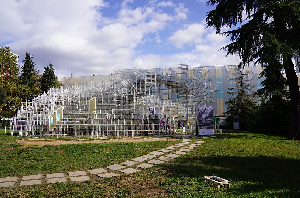 Modern metal structure with books in green park, under cloudy blue sky. Visitors walk along stepping stones. Building in background.