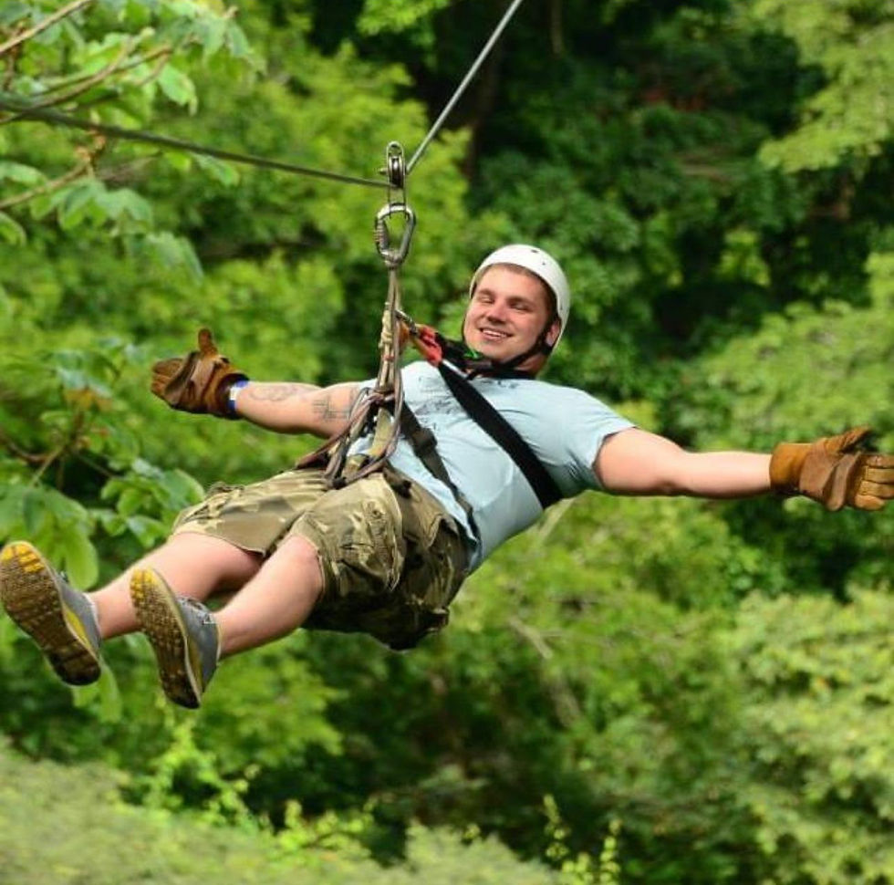 Man zip-lining through lush green forest, wearing a helmet, blue shirt, and camo shorts, smiling with arms outstretched, showing joy.