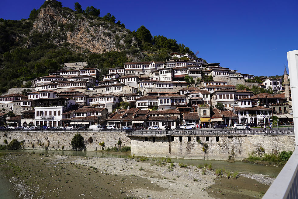 Historic hillside town with white Ottoman-style houses, red roofs, and lush greenery under a clear blue sky. People walk below along a riverbank.