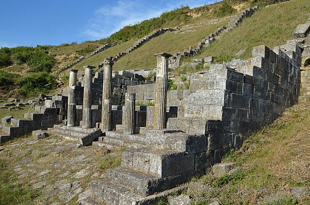 Ancient stone ruins with tall columns on grassy hillside under blue sky. The scene is serene with a sense of historical significance.