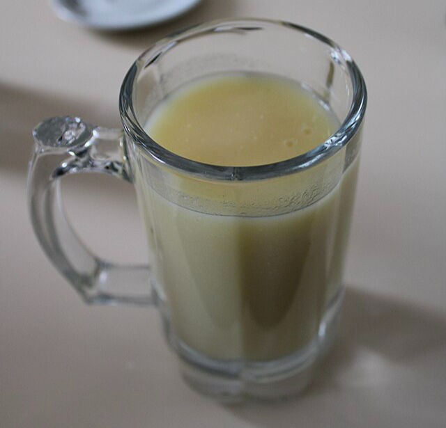 A clear mug filled with pale yellow beverage sits on a light surface. Nearby, a small white plate is partially visible, adding to a neutral mood.