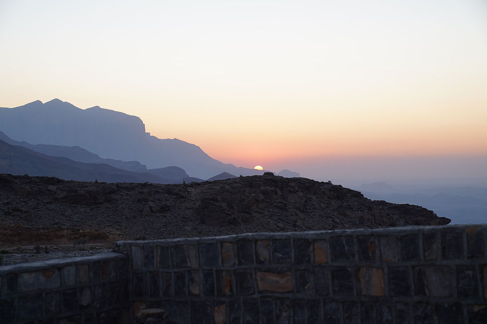 Sunset over rugged mountains with the sun partially visible. Foreground has a stone wall. Sky is a gradient of orange to blue.