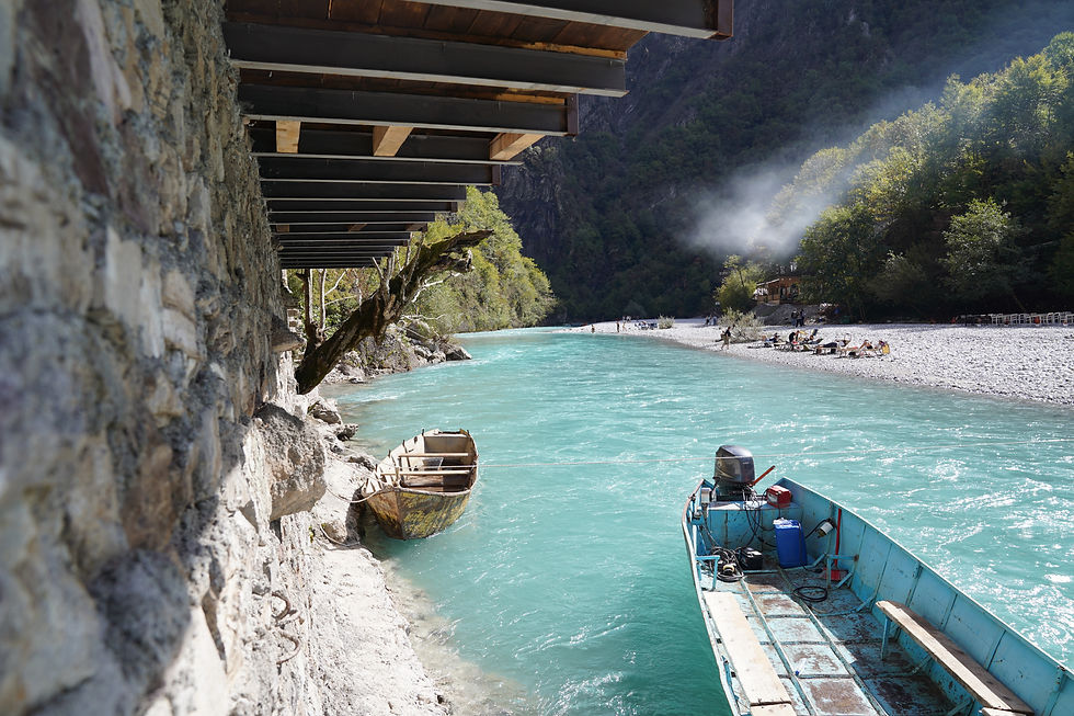 Turquoise river with boats, bordered by a stone wall. People relax on a pebbly shore under sunlit trees. Smoke drifts from a distance.
