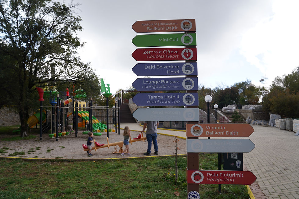 Playground scene with children on a seesaw, colorful signs pointing to restaurants and hotels, trees, and a cloudy sky in the background.