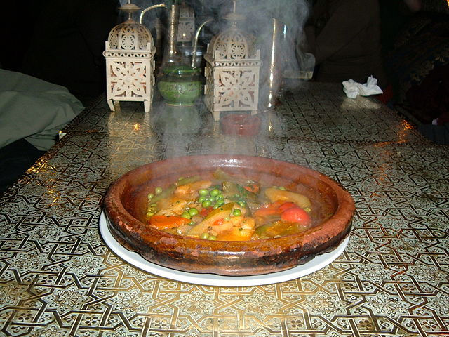 Steaming vegetable tagine on patterned table, surrounded by ornate lanterns. Warm, cozy ambiance with intricate golden designs.
