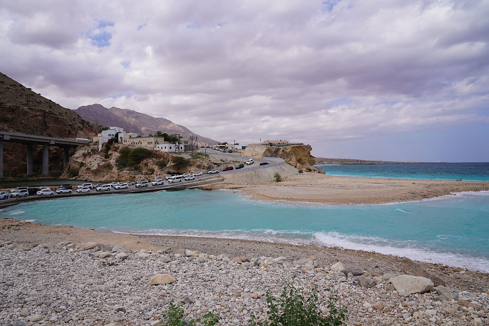 Coastal landscape with a line of cars on a road beside turquoise sea, rocky shores, and hillside houses under a cloudy sky.