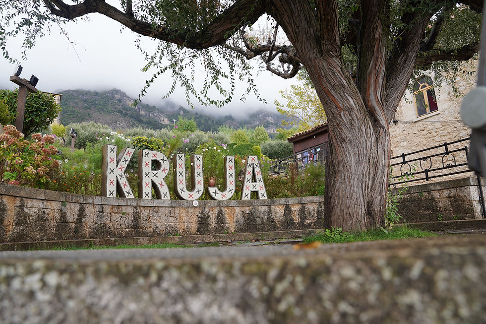 Large tree and "KRUJA" letters in a lush garden. Mountain with misty clouds in the background. Rustic stone building to the right.