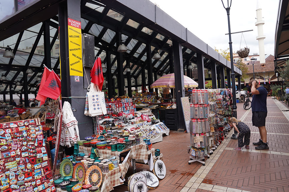 Busy outdoor market with colorful ceramics and souvenirs displayed. A man and child browse. Sign reads "Welcome" in various languages.