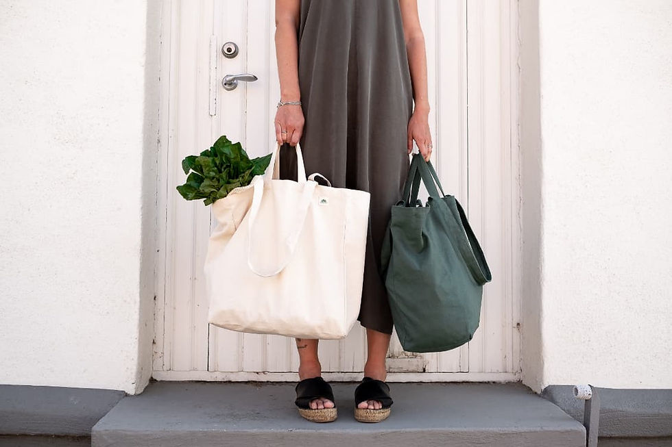 Femme tenant deux tote bags en coton bio, un naturel rempli de légumes verts et un vert sauge, devant une porte blanche – sacs de marché réutilisables zéro déchet Les Jolies Emplettes
