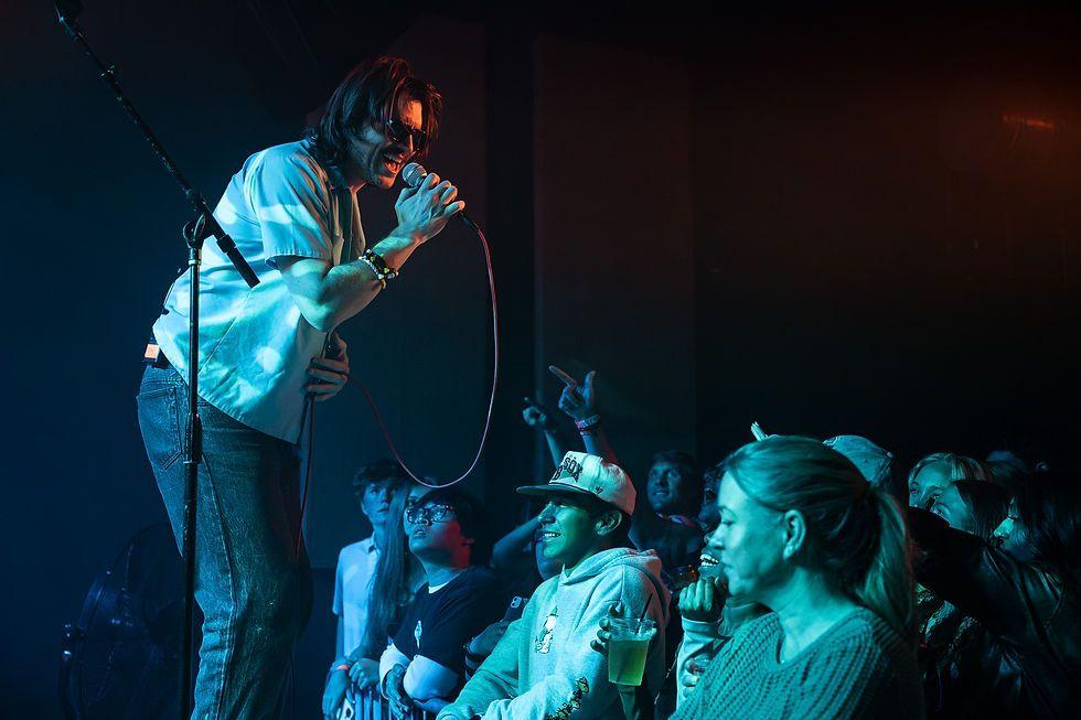 Singer in sunglasses performs energetically on stage, engaging an excited crowd in a dimly lit venue with blue and red lighting.