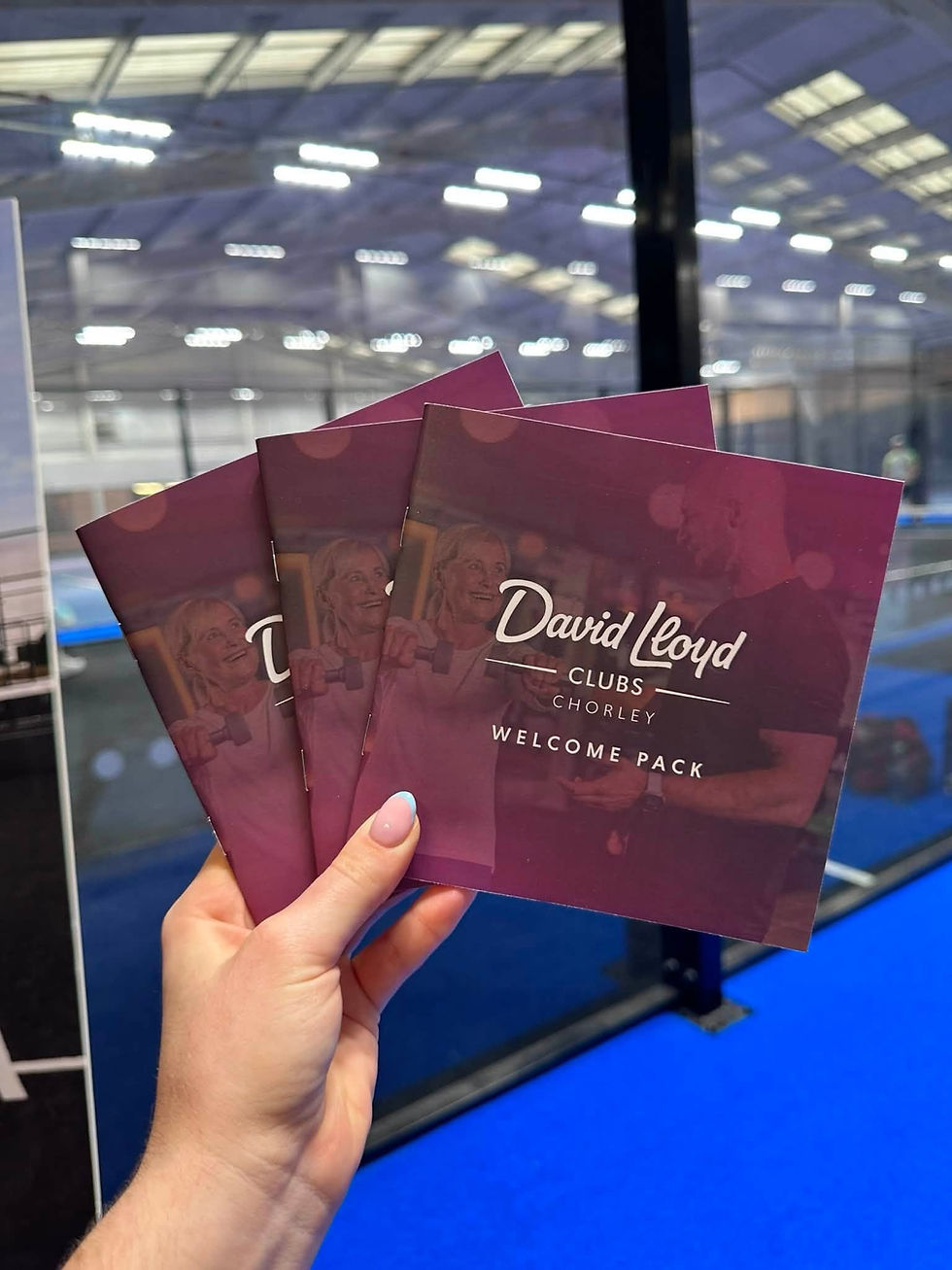 Hand holding three David Lloyd Clubs Chorley welcome packs in an indoor sports hall with blue flooring and bright lights.