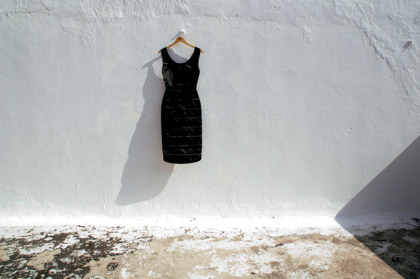 A solitary black dress hangs on a whitewashed exterior wall, casting a long shadow and questioning whether home is defined by