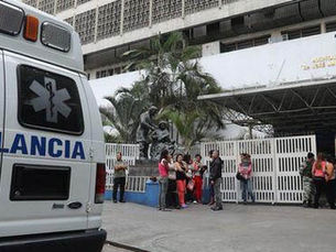 Ambulancia estacionada frente a un edificio blanco. Personas esperan ante la entrada. Ambiente urbano y tranquilo, vegetación cercana.