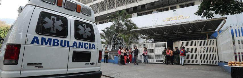 Ambulancia estacionada frente a un edificio blanco. Personas esperan ante la entrada. Ambiente urbano y tranquilo, vegetación cercana.
