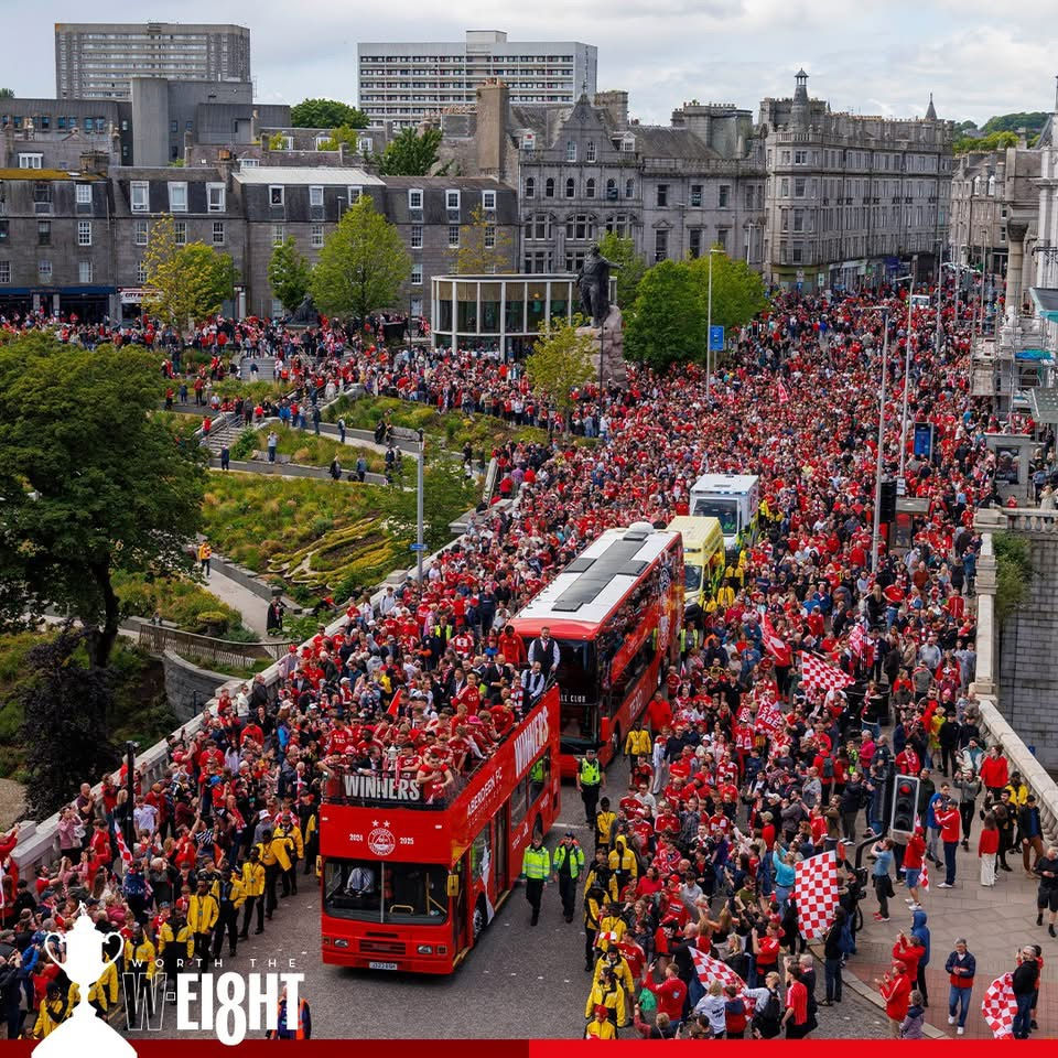 A red open top bus, surrounded by fans dressed in red with a backdrop of the grey granite buildings of Aberdeen