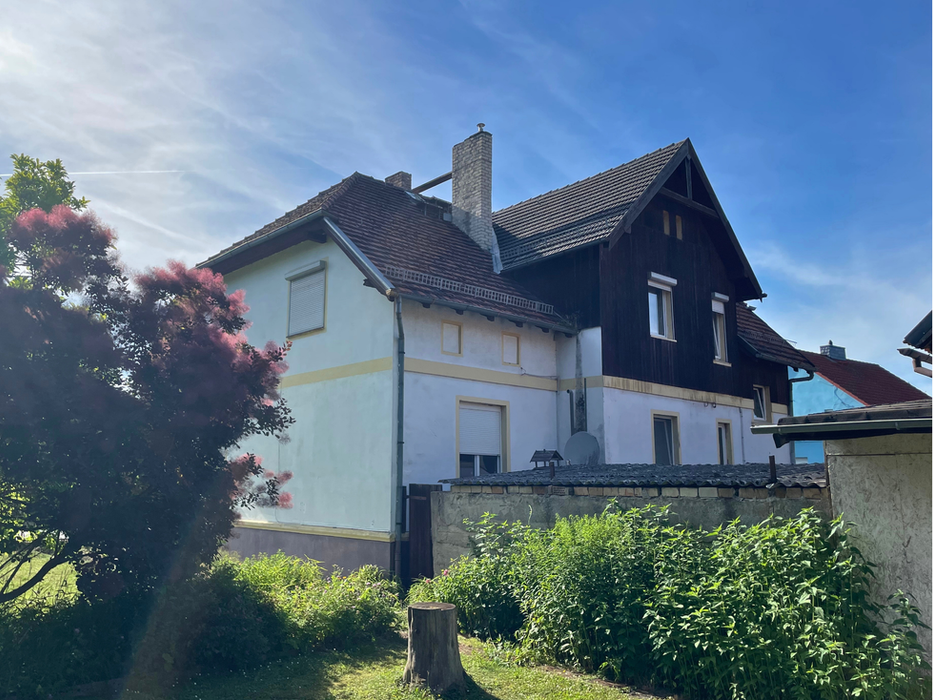Zweifamilienhaus in Nauen: Außenansicht des zweigeschossigen Hauses mit hell verputzter Fassade, dunkler Holzverkleidung im Giebelbereich und rotem Ziegeldach, umgeben von einem gründen Garten mit rosa Perückenstrauch.