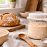 Fresh sourdough loaf, bubbly starter jar, flour, and salt arranged on a sunlit kitchen counter in a warm home kitchen.