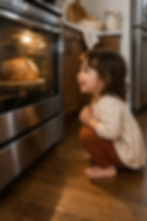 Brunette toddler girl squatting in front of the oven and smiling as she watches a loaf of sourdough baking through the oven window in a warm home kitchen.