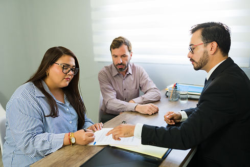 Woman signing legal agreement in a lawyer's office, husband sitting next to her during a c