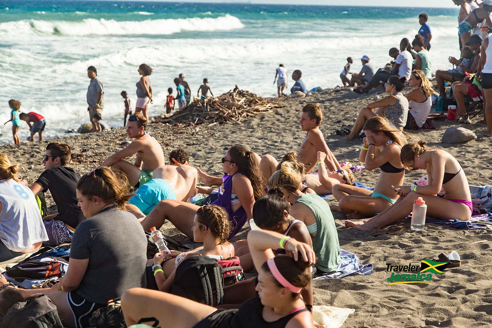 Beachgoers relax and enjoy a sunny day by the waves, sprawled across the sandy shores, while others play and walk along the water's edge at Pali Pro surf Competition Jamaica.