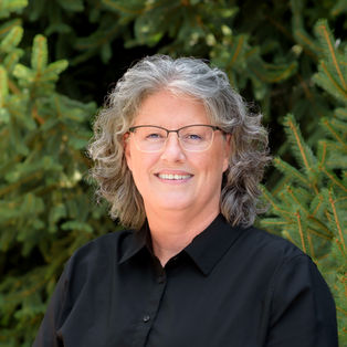 Professional headshot of an older woman with curly gray hair wearing glasses and a black blouse, standing outdoors in front of green trees.