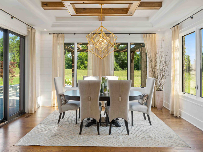 Luxury custom dining room by Frazier Homes featuring floor-to-ceiling windows, a coffered ceiling with wood beam accents, designer geometric chandelier, elegant upholstered dining chairs, and seamless indoor-outdoor views.
