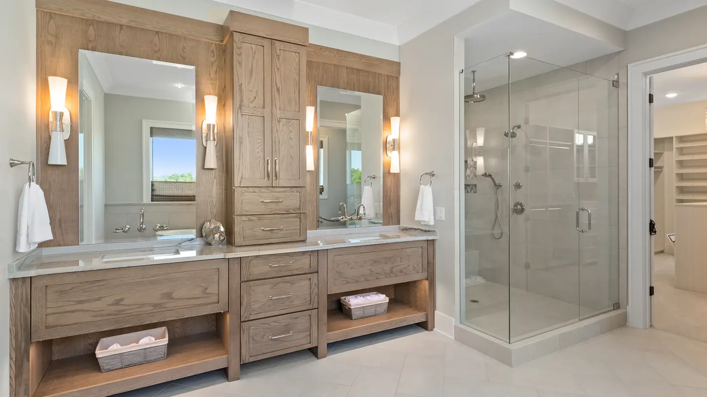 Luxury master bathroom featuring custom wood double vanities with stone countertops, modern sconces, spacious glass shower with multiple fixtures, and soft neutral tile flooring in a Frazier Homes Homearama-winning design.