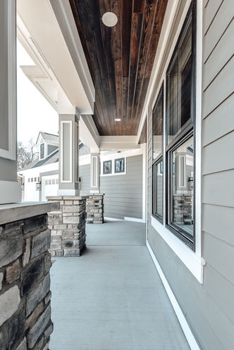 A stunning covered porch by Frazier Homes, showcasing stone columns, a rich wooden ceiling, and paneled architectural details. The large windows and meticulously crafted design elements enhance the inviting atmosphere, making this outdoor space perfect for relaxation and entertaining. Experience the superior craftsmanship and timeless beauty that Frazier Homes brings to every project.