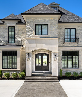 Front elevation of a luxury custom home by Frazier Homes featuring elegant stonework, black-framed windows, wrought iron balconies, and a grand arched double-door entry with gas lanterns and landscaped walkway.