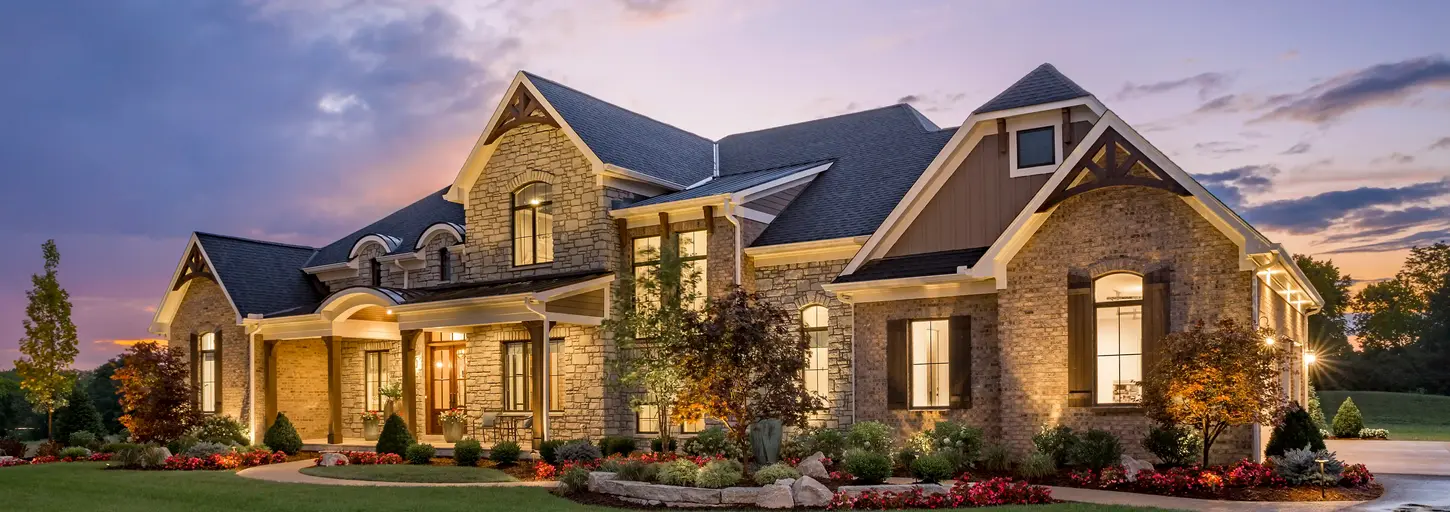 Exterior view of a modern home, winner of Homearama 2018, with wood detailing and lush landscaping. The setting sun casts a warm glow, accentuating the home's contemporary design.