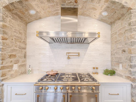 Luxury custom kitchen by Frazier Homes featuring a professional gas range, stone arch surround, white tile backsplash, and gold-accented fixtures, showcasing craftsmanship and attention to detail by an experienced general contractor.