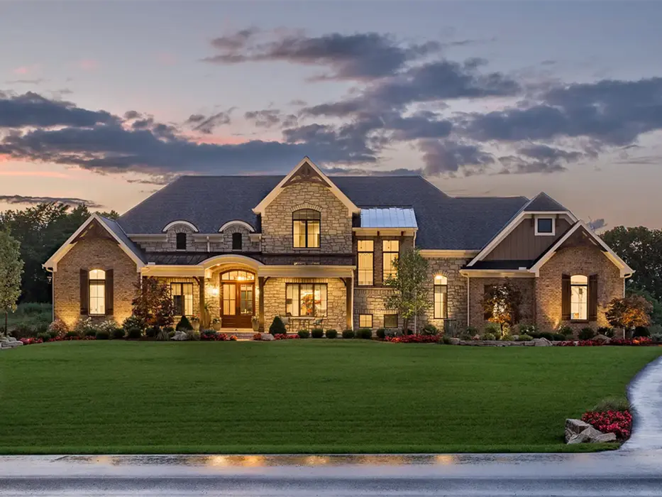 Exterior view of a modern home, winner of Homearama 2018, with wood detailing and lush landscaping. The setting sun casts a warm glow, accentuating the home's contemporary design.