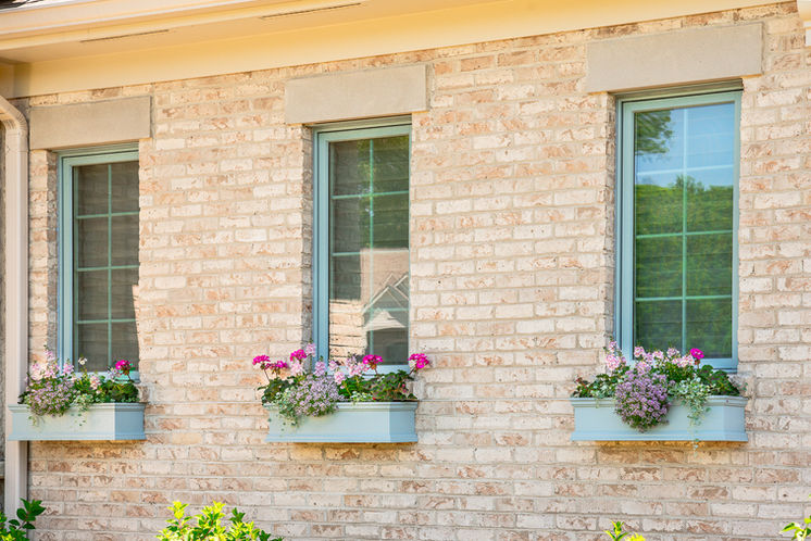 Three windows with soft blue trim and flower boxes filled with pink and purple blooms on a light brick home.