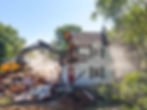 Excavator tearing down an existing residential home during demolition phase in preparation for a custom home rebuild, with debris, exposed structure, and surrounding trees visible on site.