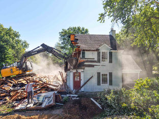 Excavator tearing down an existing residential home during demolition phase in preparation for a custom home rebuild, with debris, exposed structure, and surrounding trees visible on site.