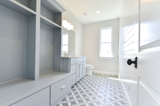 Bathroom featuring built-in blue cabinetry with drawers and open shelving, a light wood vanity with chrome fixtures, geometric gray-and-white patterned tile flooring, white shiplap door, and two tall windows letting in natural light.