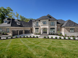 Elegant custom-built home by Frazier Homes featuring stone and brick exterior, black three-car garage doors, and beautifully landscaped front yard under a clear blue sky.